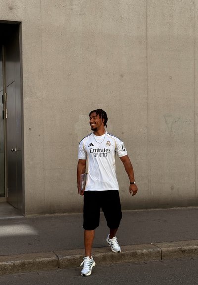 Hombre caminando por la acera cerca de una pared de hormigón vistiendo una camiseta blanca del Real Madrid, pantalones cortos negros, zapatillas blancas, sosteniendo un libro o carpeta.