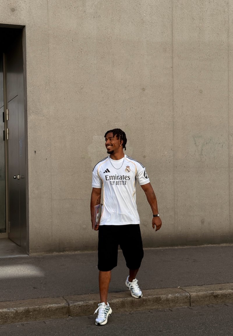 Uomo che cammina sul marciapiede vicino a un muro di cemento, indossa una maglietta bianca del Real Madrid, pantaloncini neri, scarpe da ginnastica bianche, tiene un libro o una cartella.