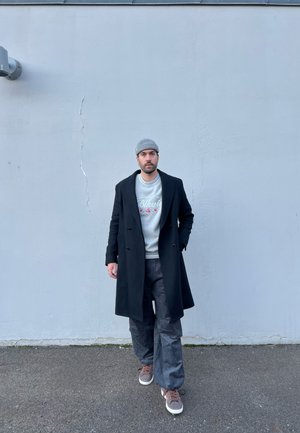 Abrigo negro de doble botonadura sobre un sudadera gris claro con texto floral, pantalones oscuros de pierna ancha y zapatillas marrones, con un fondo de pared gris.