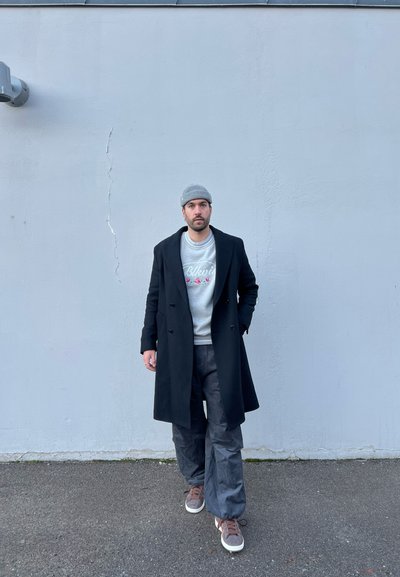 Abrigo negro de doble botonadura sobre un sudadera gris claro con texto floral, pantalones oscuros de pierna ancha y zapatillas marrones, con un fondo de pared gris.