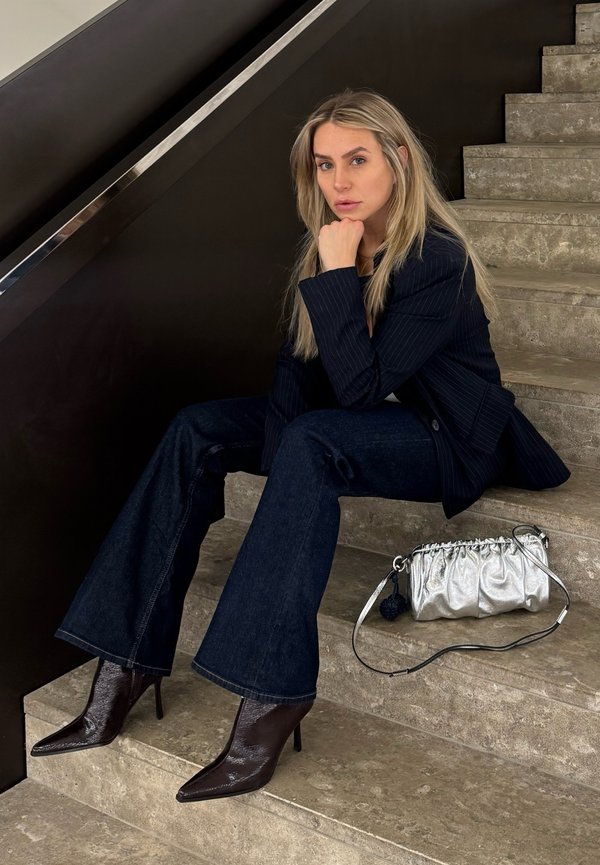 Femeie cu păr blond lung stă pe scări de piatră, purtând un sacou închis la culoare, blugi evazați, cizme cu toc ascuțit, având o geantă argintie alături.