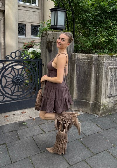 Joven mujer con un vestido sin mangas marrón y botas altas con flecos posa sonriendo cerca de una puerta de hierro forjado y una pared de piedra con vegetación.
