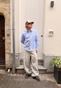 Šviesiai mėlyna marškiniai su sagomis, pilkos sportinės kelnės, rudi batai; stovi prie tekstūruotos akmens sienos, netoli balti durys ir žaluma.