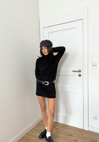 Robe-pull noire à col roulé avec une ceinture marron et une boucle argentée, associée à des mocassins noirs et une casquette grise, sur un fond de mur neutre.