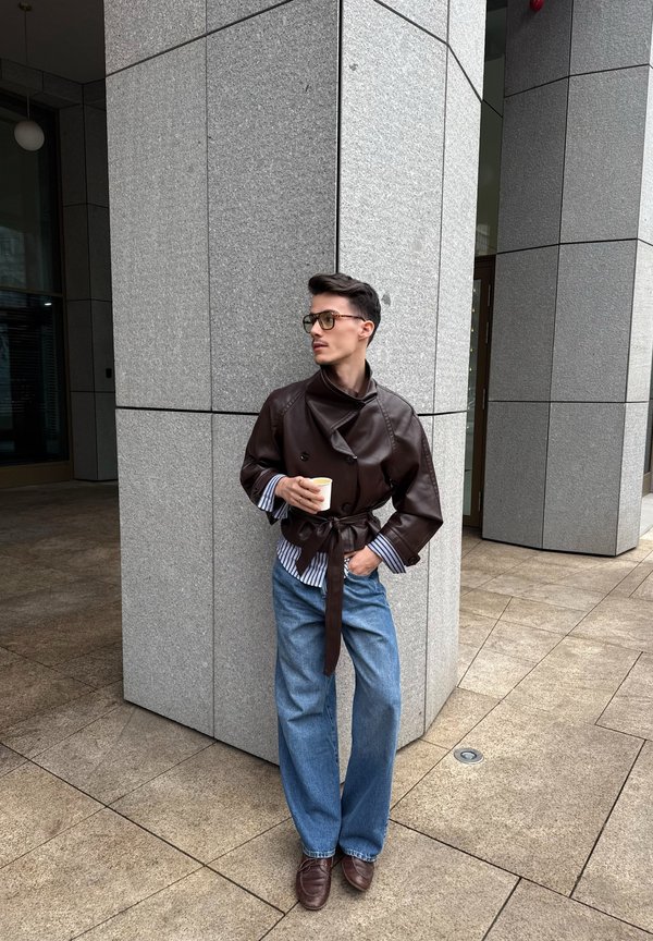 Person trägt eine braune Lederjacke, gestreiftes Hemd, blaue Jeans und Brille, hält eine Tasse und lehnt sich im Freien an eine große graue Steinsäule.