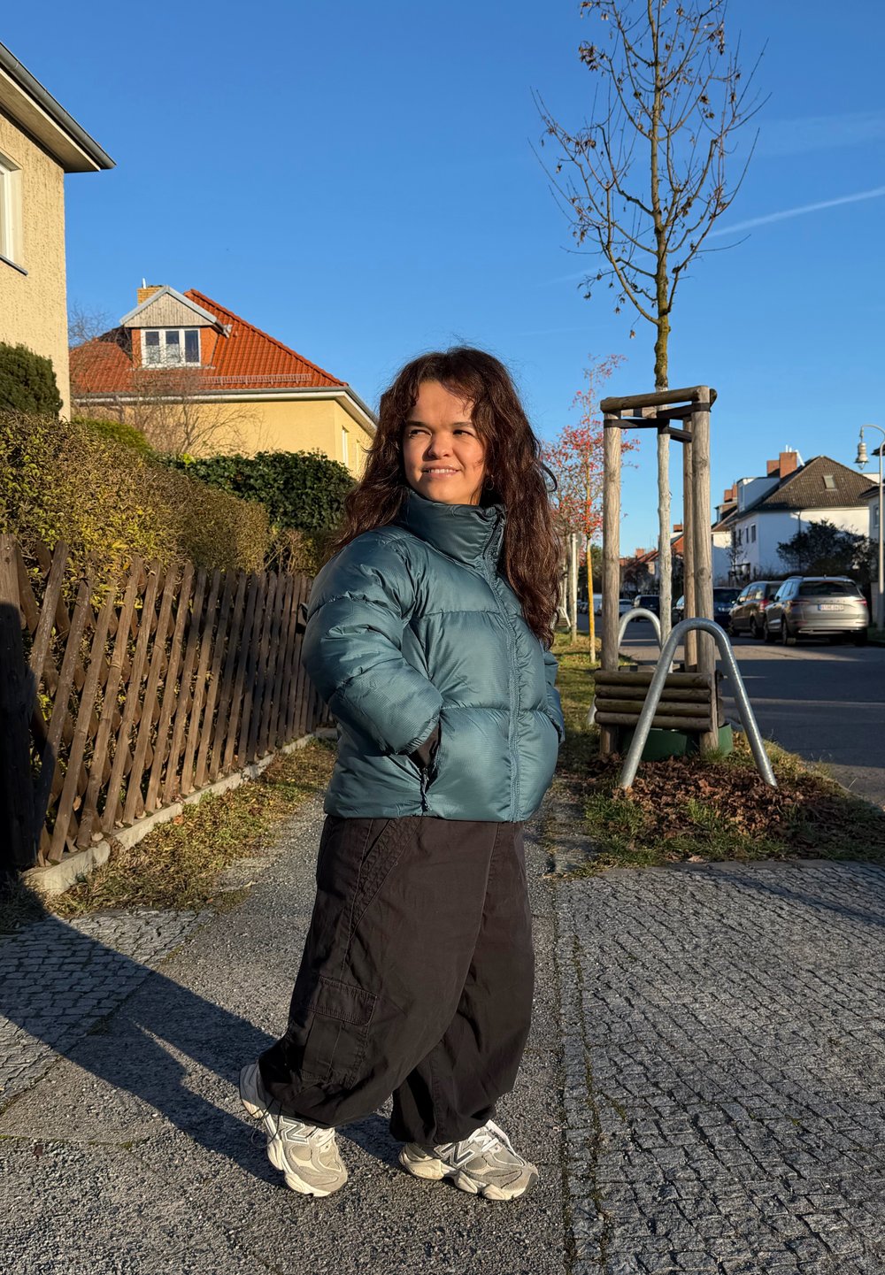 Jeune femme portant une veste bleu sarcelle et un pantalon noir marchant sur un trottoir pavé près de maisons résidentielles sous un ciel bleu clair.