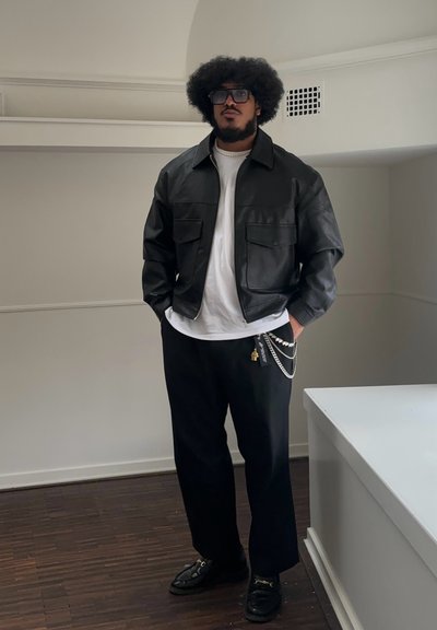 Chaqueta de cuero negra con dos bolsillos frontales, camiseta blanca, pantalones holgados negros, accesorio de cadenas en capas y mocasines negros sobre un suelo de madera.