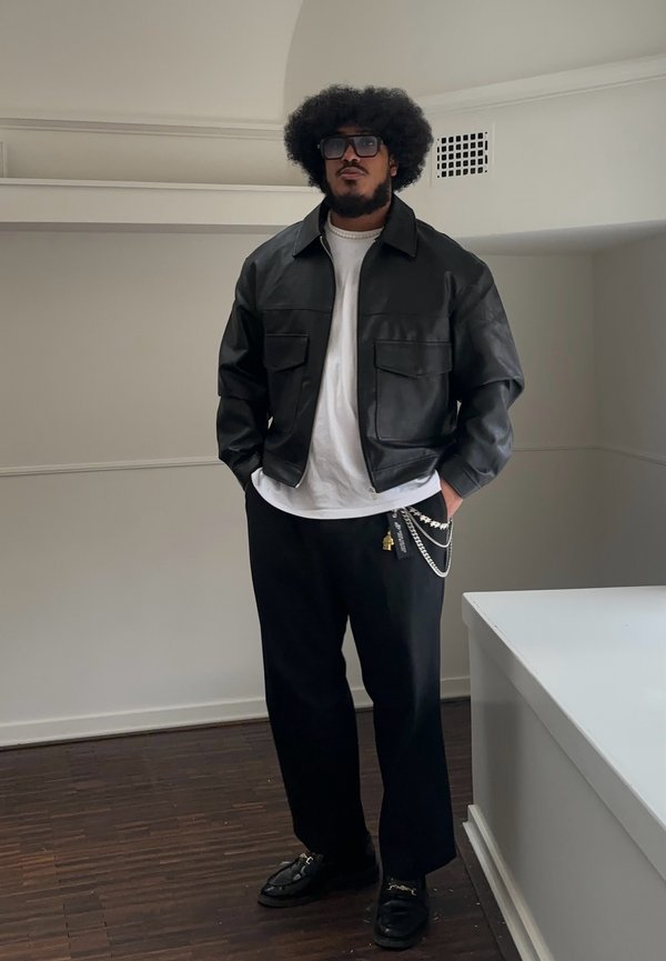 Black leather jacket with two front pockets, white t-shirt, loose black trousers, layered chain accessory, and black loafers on a wooden floor.