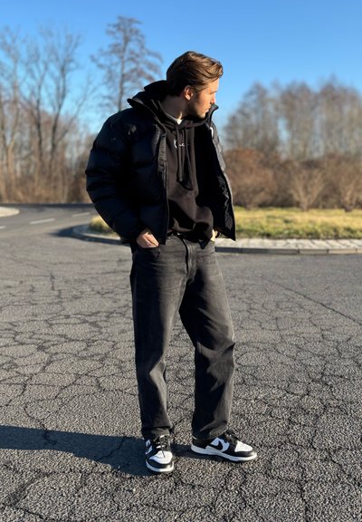 Chaqueta de plumas negra sobre un sudadera negra, jeans grises y zapatillas en blanco y negro. El escenario es un área pavimentada al aire libre con algunos árboles dispersos.