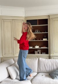 Mujer con un suéter rojo y jeans azules bailando descalza sobre un sofá beige en una sala de estar con estanterías y armarios empotrados.