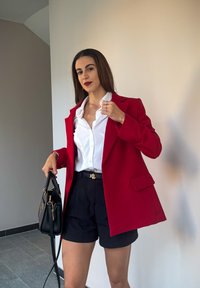 Rød skræddersyet blazer over en hvid skjorte med knapper, parret med sorte shorts og en sort håndtaske. Har gulddetaljer på bæltet.