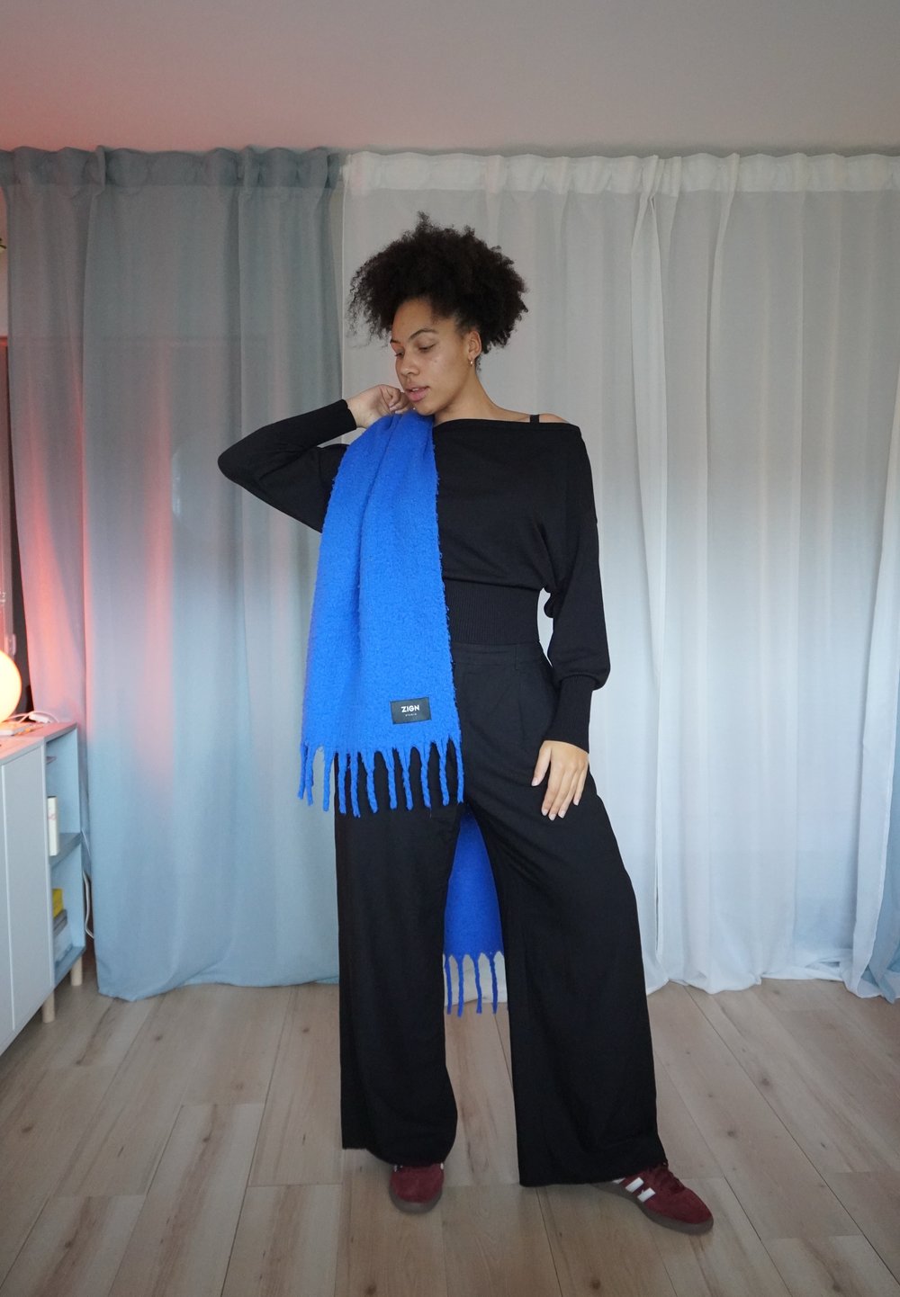 Svart off-shoulder topp, løstsittende svarte bukser og et skarpt blått skjerf med frynser og en tagg. Myk tekstur; minimalistisk design.