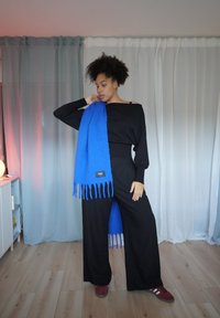 Svart off-shoulder topp, løstsittende svarte bukser og et skarpt blått skjerf med frynser og en tagg. Myk tekstur; minimalistisk design.