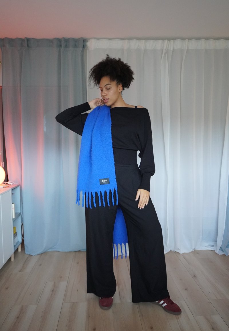Top preto ombro a ombro, calças pretas largas e lenço azul brilhante com franjas e etiqueta. Textura suave; design minimalista.