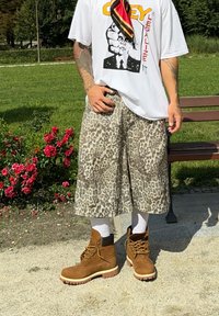 Vékony anyagból készült leopárdmintás shorts, barna bokacsizmával és fehér zoknival párosítva. Fehér póló grafikus mintával.