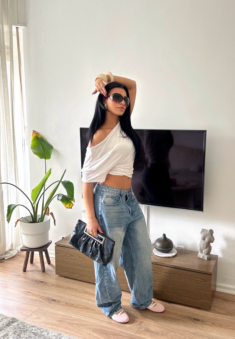 Femme posant à l'intérieur vêtue d'un jean oversize, d'un top blanc à épaules dénudées, lunettes de soleil, tenant une pochette noire, debout près d'une télévision et d'une plante.