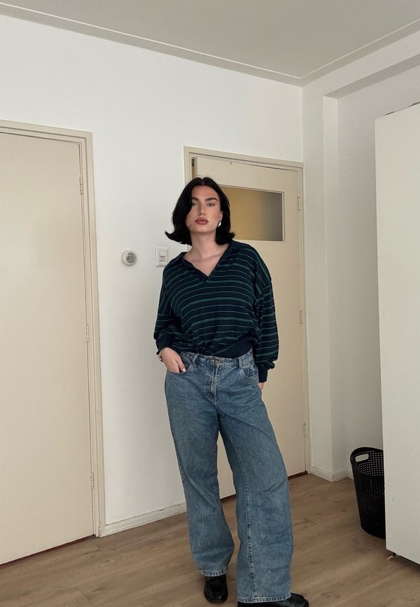 Fekete hajú nő áll bent, sötét csíkos hosszú ujjú inget, bő kék farmert és fekete cipőt visel, kezét zsebre dugva.