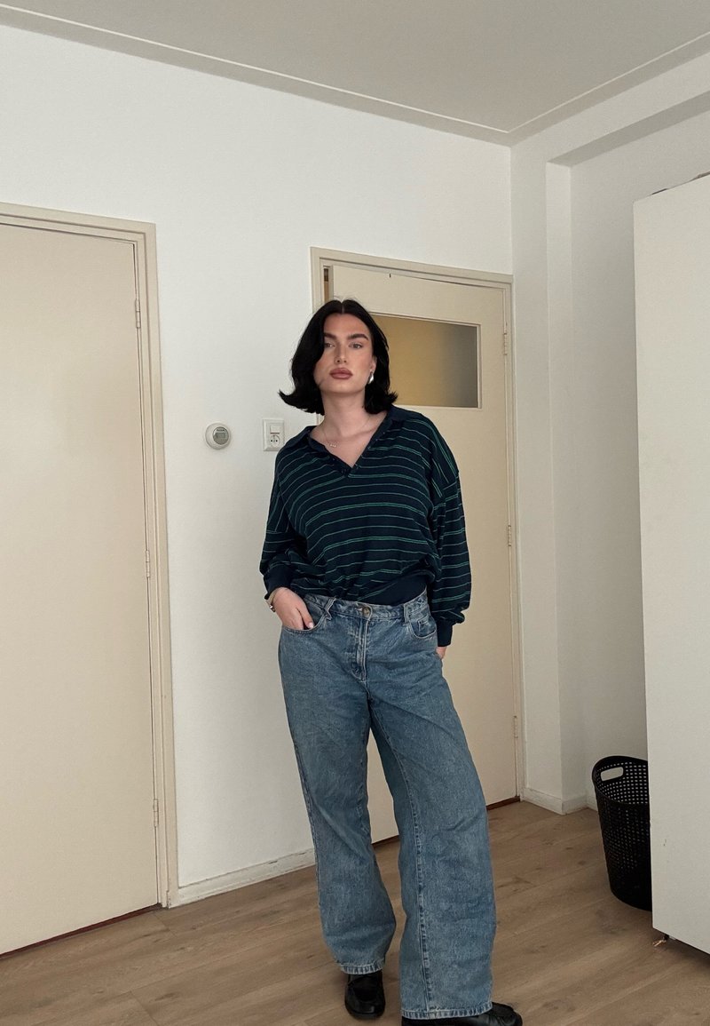 Femme aux cheveux noirs debout à l'intérieur, portant une chemise à manches longues rayée foncée, un jean bleu ample et des chaussures noires, les mains dans les poches.