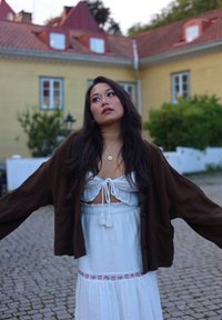 Bruine, korte linnen jas over een witte, zachtvallende jurk met trekkoord in de taille en bloemrijke borduursels. Op de achtergrond een geel gebouw.