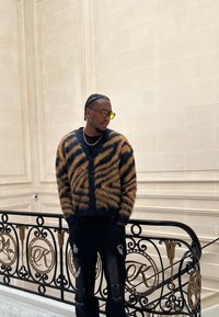 Cardigan texturé avec des rayures orange et bleu foncé, fermeture à boutons, porté sur une chemise noire ; associé à un jean noir déchiré.