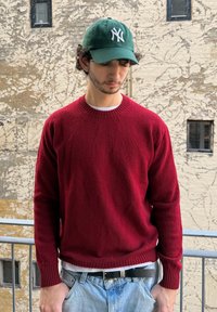 Punane villane kampsun soonikkoega krae ja mansettidega, kantud valge särgi kohal. Hele sinine teksapüksid. Roheline nokamüts valge logoga.