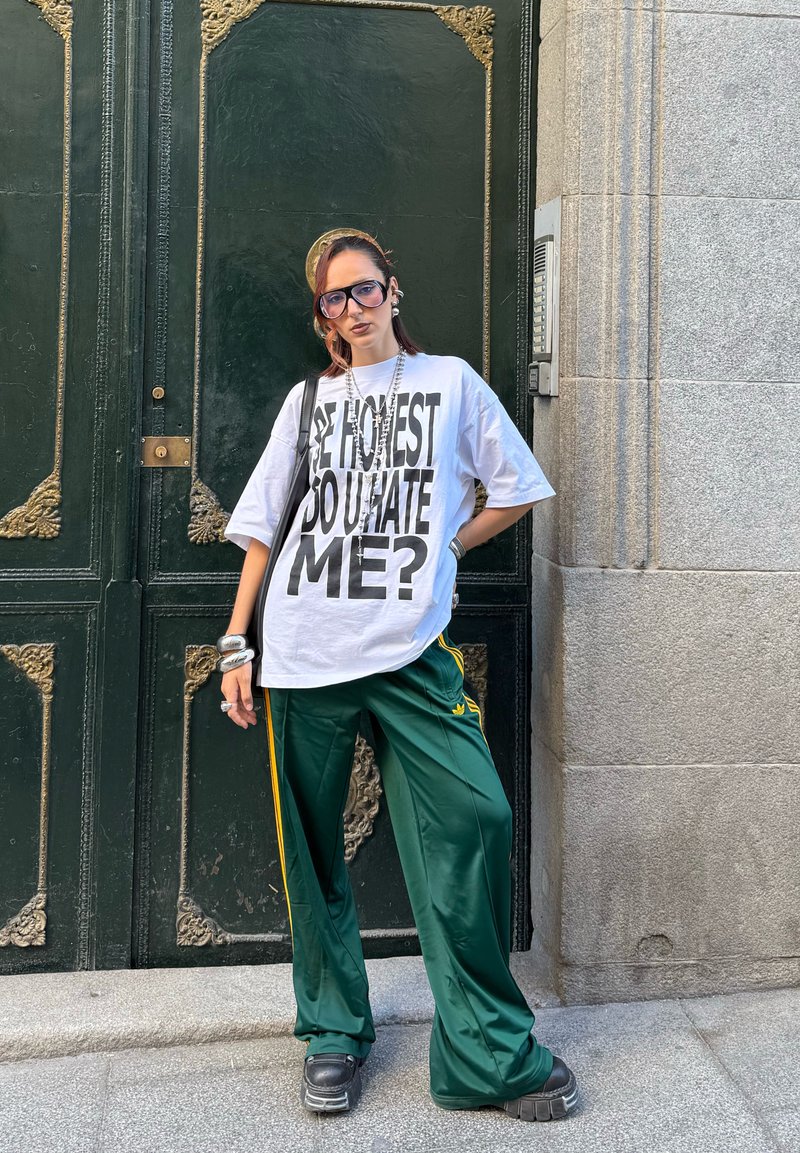 Personne portant de grandes lunettes, une chemise blanche surdimensionnée avec le texte "SOIS HONNÊTE, ME HAIS-TU ?", un pantalon de survêtement vert et des grosses chaussures noires, se tenant devant une porte ornée de couleur vert foncé.