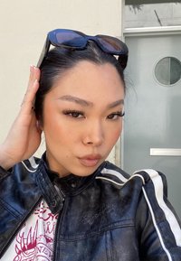 Veste en cuir noire avec des rayures blanches, portée sur un t-shirt graphique. Des lunettes de soleil reposent sur le sommet de la tête. Fond clair avec une porte en verre.