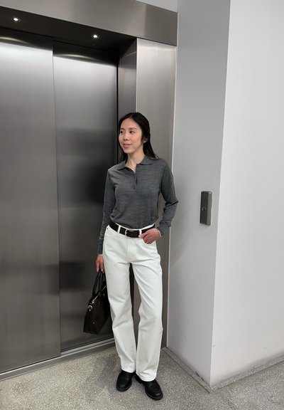 Mujer con camisa gris de manga larga y pantalones blancos está junto a unas puertas de ascensor de acero inoxidable cerradas sosteniendo un bolso negro.