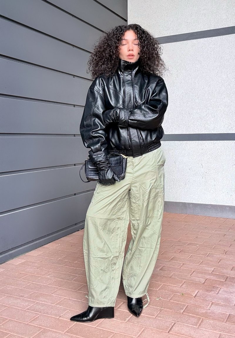 Black leather bomber jacket with a high collar, paired with light green oversized pants. Black square-toed ankle boots and a textured black clutch.