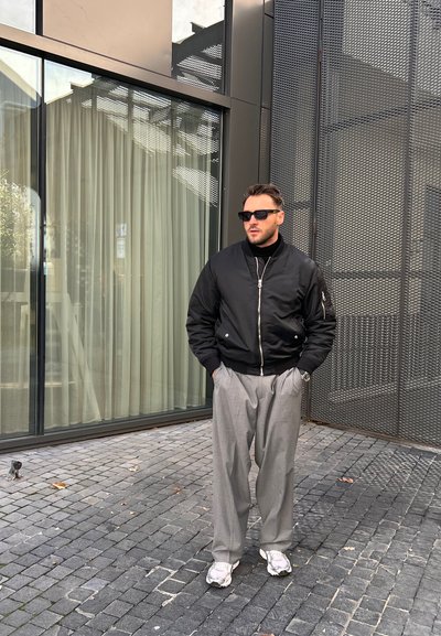 Chaqueta bomber negra con cremallera frontal, pantalones gris con pliegues y zapatillas blancas. Los accesorios incluyen gafas de sol y un reloj.