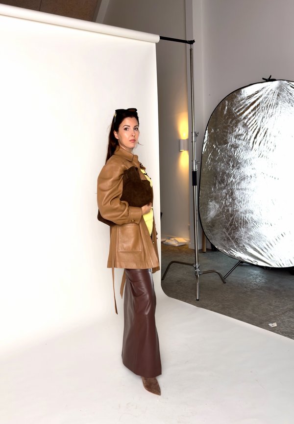 Une femme pose dans un manteau en cuir beige et une jupe brun foncé sur le plateau d’un studio photo, à côté d’un grand réflecteur argenté.