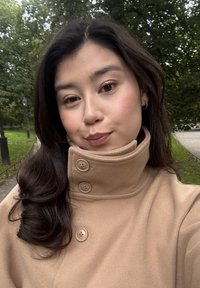 Ung kvinna med långt mörkt hår som bär en knäppt beige kappa utomhus i en park med träd och en gångväg.