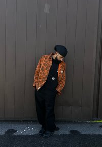 Πορτοκαλί πουκάμισο με παραλλαγή πάνω από μαύρο τζάκετ, σε συνδυασμό με μαύρες σαλοπέτες και μαύρα παπούτσια, μπροστά από καφέ ξύλινο φόντο.