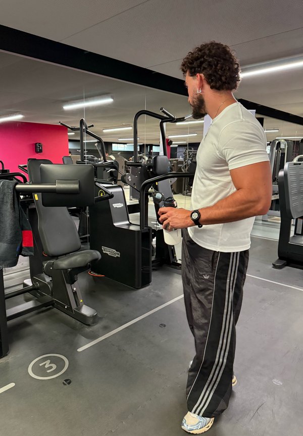 Mann in weißem Hemd hält Wasserflasche und steht neben Fitnessgeräten in einem Fitnessstudio mit Spiegeln.