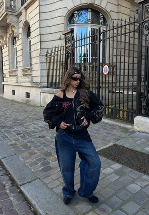Joven con chaqueta negra extragrande, vaqueros anchos, gafas de sol y diadema, está de pie en una calle empedrada frente a una valla de hierro ornamentada y un edificio de piedra.