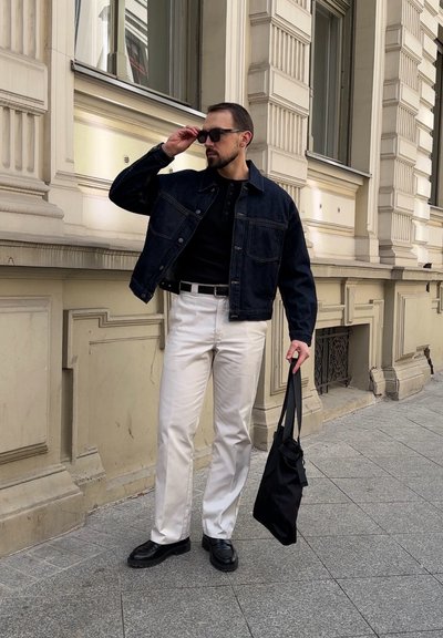 Hombre con gafas de sol negras, chaqueta vaquera oscura, camisa negra, pantalones blancos, sosteniendo un bolso tote negro, de pie en la acera de la ciudad junto a un edificio ornamentado.