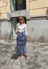 Λευκό πουκάμισο με κουμπιά και μακριά μανίκια, μίντι φούστα με μπλε λουλουδάτο σχέδιο, λευκές σαγιονάρες, χρυσή τσάντα αλυσίδας και γυαλιά ηλίου. Φωτεινός εξωτερικός χώρος.