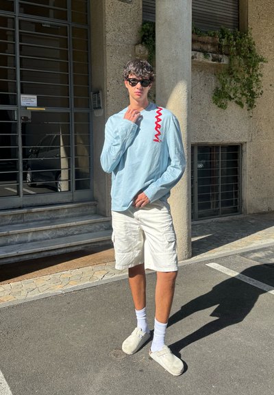 Camisa de manga larga azul claro con un patrón de zigzag rojo, pantalones cortos cargo blancos, zapatos gris oscuro sin cordones y calcetines blancos, de pie al aire libre.