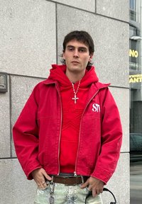 Νεαρός άνδρας που φοράει κόκκινο μπουφάν και φούτερ με σχέδιο άστρου, standing against a gray concrete wall holding headphones in one hand.