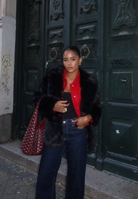 Palton din blană neagr ă peste o cămașă roșie cu nasturi, asortat cu blugi închiși la culoare cu talie înaltă și o geantă roșie cu studuri. Fond cu uși verzi.