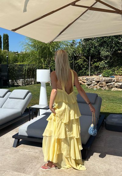 Vestido amarillo sin mangas, de capas con volantes, de tela ligera. Se lleva con un pequeño bolso azul, en un patio con tumbonas.