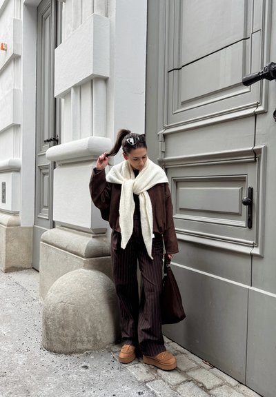 Mujer con coleta y gafas de sol sostiene su cabello, vestida con chaqueta marrón, bufanda crema, pantalones de rayas finas y zapatos beige, junto a un edificio gris.