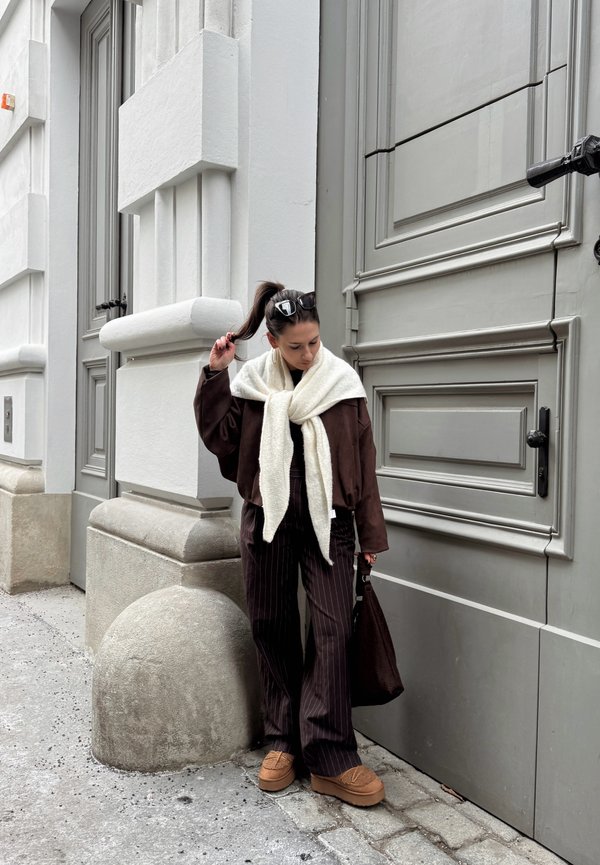 Femme avec queue de cheval et lunettes de soleil tenant ses cheveux, portant une veste marron, une écharpe crème, un pantalon à rayures fines et des chaussures beiges, debout près d'un bâtiment gris.