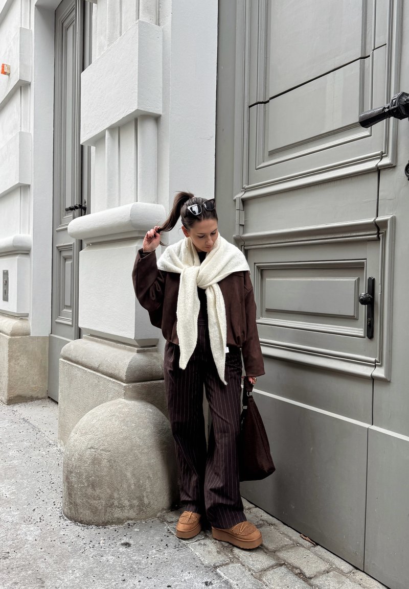 Kobieta z kucykiem i okularami przeciwsłonecznymi trzyma włosy, ubrana w brązową kurtkę, kremowy szalik, spodnie w cienkie paski i beżowe buty, stoi przy szarym budynku.