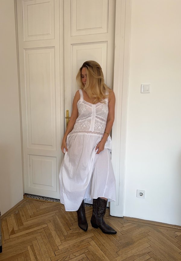 Abito bianco con dettagli in pizzo e spalline regolabili, abbinato a stivali da cowboy marrone scuro. Sullo sfondo si vede un pavimento in legno e una porta bianca.