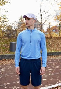 Gaiši zils sportisks tops ar garām piedurknēm un pusgaru rāvējslēdzēju, kopā ar tumši ziliem šortiem. Audumam ir faktūras raksts, kas piemērots āra aktivitātēm.