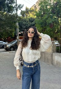 Blouse crème transparente à manches bouffantes, associée à un jean en denim bleu taille haute et une ceinture chaîne dorée décorative. Sac à épaule à motifs.