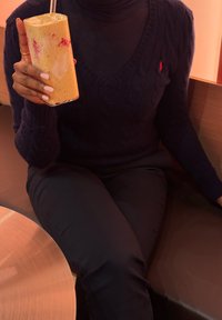 Smoothie korkeassa lasissa, vaaleankeltainen pinkeillä kierroksilla. Henkilö laivastonsinisessä kaapelineuleessa ja tummissa housuissa, istumassa puupöydän ääressä.