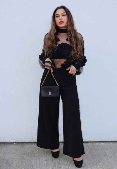 Blusa negra transparente con detalles florales, combinada con pantalones negros de pierna ancha y un pequeño bolso negro con cadena dorada.