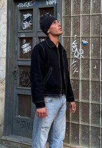 Uomo con giacca nera, berretto nero e jeans azzurri strappati che guarda verso l'alto accanto a una porta coperta di graffiti e blocchi di vetro.